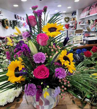 Carnival - A vibrant and uplifting arrangement designed to celebrate a life filled with warmth, happiness, and treasured memories. Featuring a striking blend of sunny yellow, rich pink, and deep purple tones with elegant greenery, this tribute brings comfort and brightness during difficult times. Presented in a contemporary keepsake container, it offers a heartfelt expression of love, remembrance, and gratitude for a dearly missed loved one.