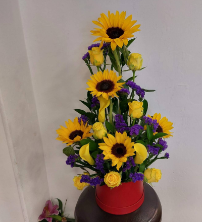 Sunflower Hatbox - A vibrant and uplifting arrangement designed in a striking upright style, featuring radiant golden tones contrasted with rich purple accents and lush greenery. Presented in a bold red hatbox, this design brings height, elegance, and cheerful energy to any setting. Perfect for celebrations, birthdays, thank-you gifts, or brightening someone’s day with a stunning burst of colour.
