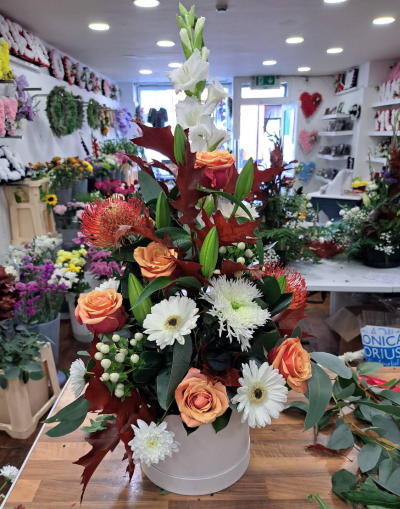 Autumn Elegance - A sophisticated tall arrangement inspired by rich autumn tones and natural textures. Featuring warm peach shades, crisp whites, and deep seasonal foliage, this striking design brings height, movement, and elegance to any room. Presented in a modern hatbox container, it makes a beautiful statement gift for celebrations, special occasions, or stylish home décor.
